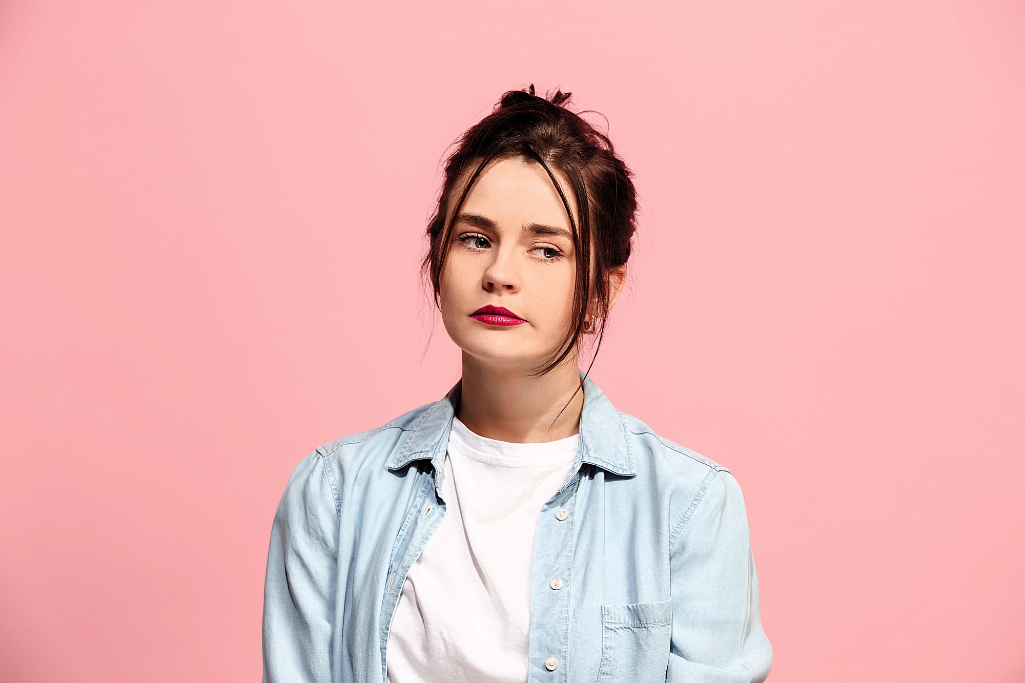 Woman with dark hair in a bun, wearing a light denim shirt over a white t-shirt, standing against a pink background, looking to the side with a neutral expression.