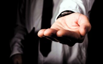 A person in a shirt and tie extends their open hand forward, palm up, against a dark background. The gesture appears inviting or offering assistance.