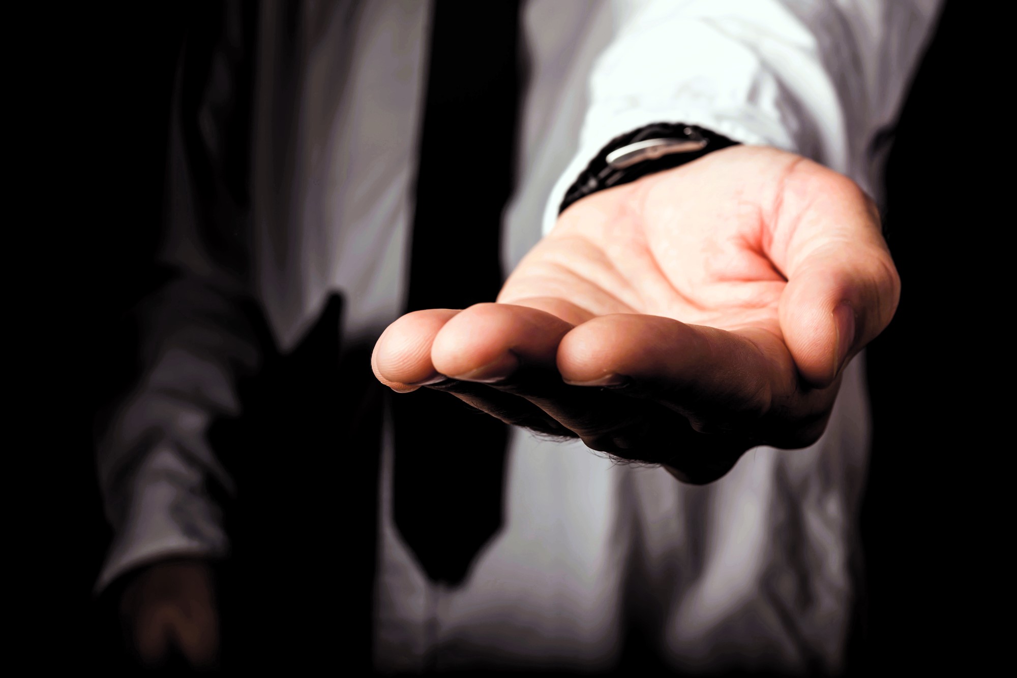 A person in a shirt and tie extends their open hand forward, palm up, against a dark background. The gesture appears inviting or offering assistance.