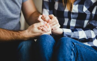 A close-up of two people sitting closely. One is wearing a gray shirt, and the other is in a plaid shirt and jeans. They are holding hands, conveying comfort and support.