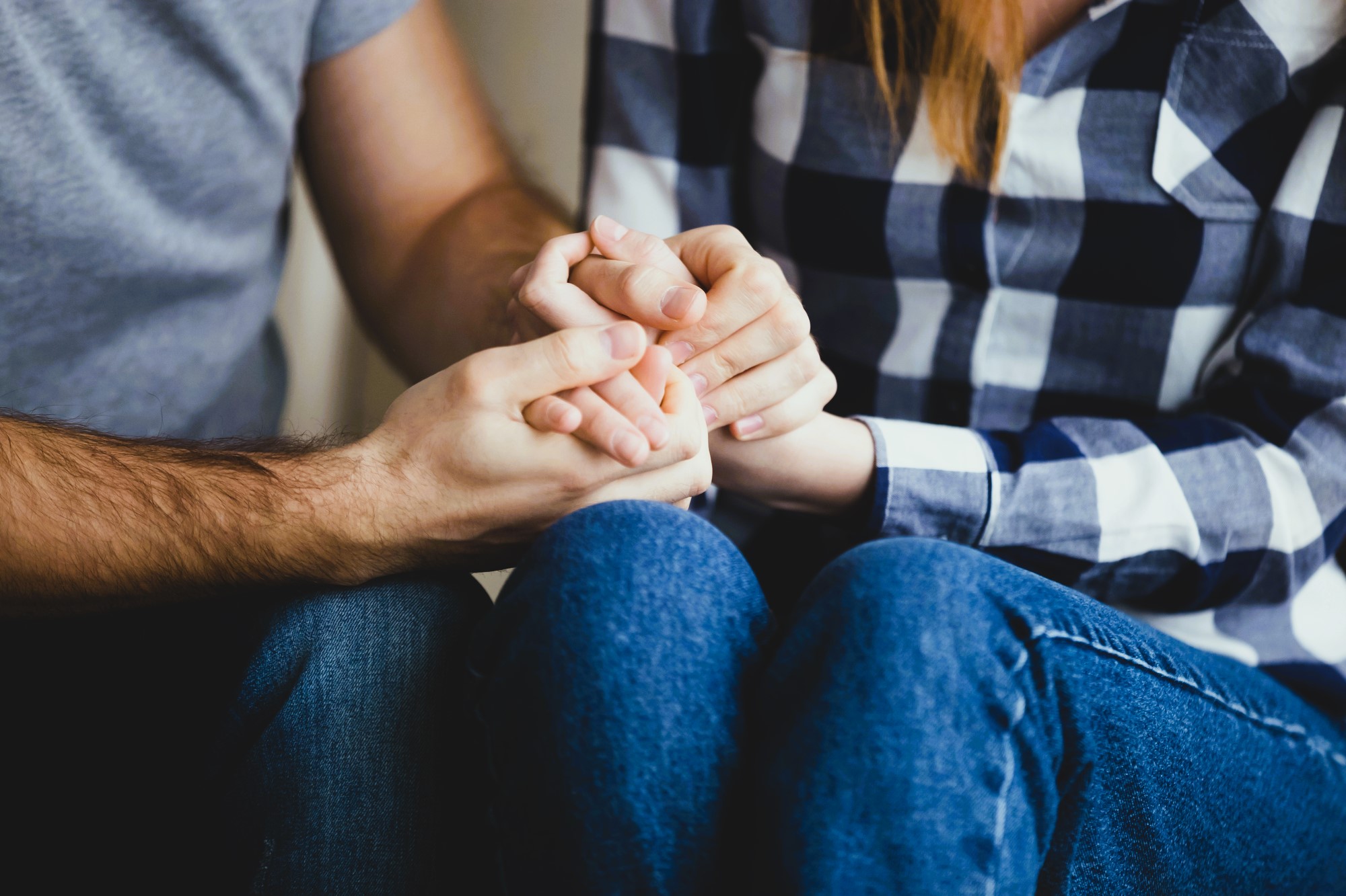 A close-up of two people sitting closely. One is wearing a gray shirt, and the other is in a plaid shirt and jeans. They are holding hands, conveying comfort and support.