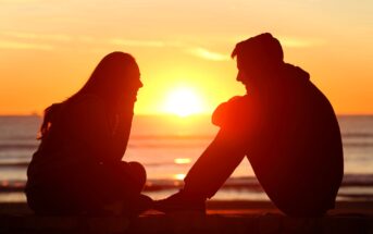 Silhouetted against a vibrant sunset, two people sit facing each other by the ocean. The sun is setting on the horizon, casting an orange glow over the scene. One person appears to be speaking, adding a sense of conversation and connection.