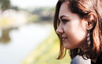 A woman with brown hair and pink flower-shaped earrings looks thoughtfully at a blurred outdoor scene, possibly a riverbank or park. The soft background highlights her contemplative mood.