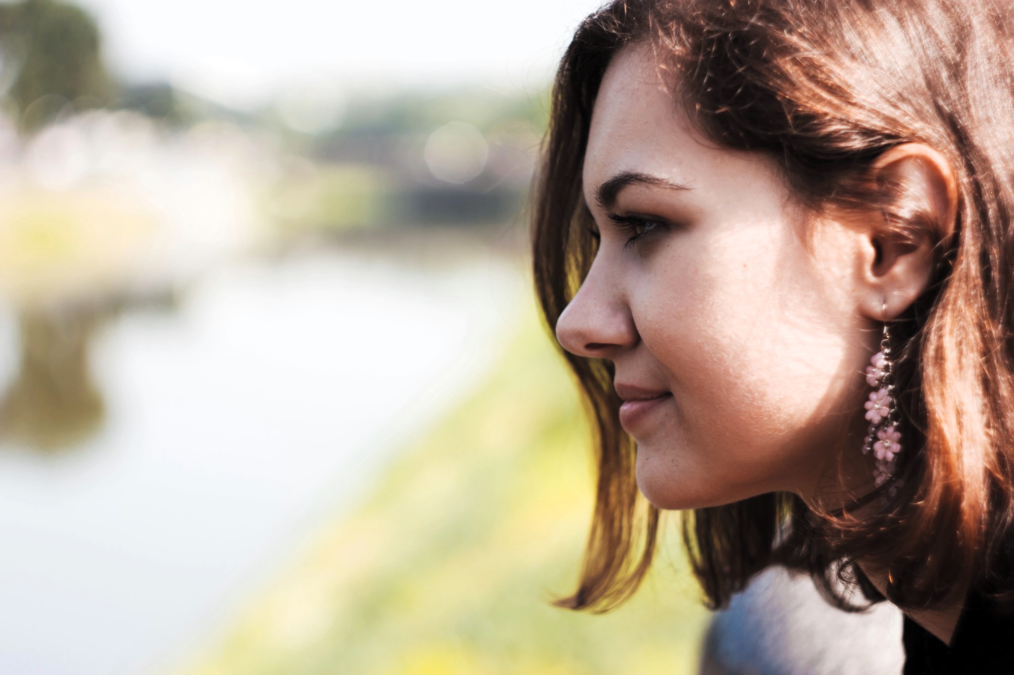 A woman with brown hair and pink flower-shaped earrings looks thoughtfully at a blurred outdoor scene, possibly a riverbank or park. The soft background highlights her contemplative mood.