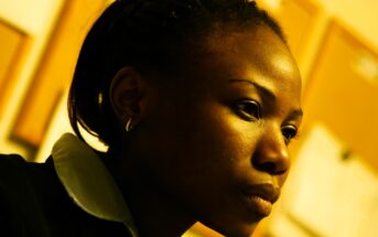 Profile of a contemplative person with braided hair and a collared shirt, set against a blurred background of bulletin boards. The warm lighting casts a soft glow on their face, highlighting their thoughtful expression.