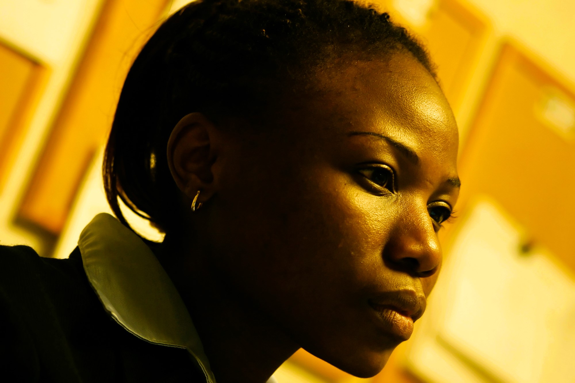 Profile of a contemplative person with braided hair and a collared shirt, set against a blurred background of bulletin boards. The warm lighting casts a soft glow on their face, highlighting their thoughtful expression.