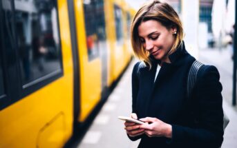 A woman with short hair is smiling while looking at her smartphone. She is standing next to a yellow train on a city street. She is wearing a black jacket and has a backpack. The background is slightly blurred.