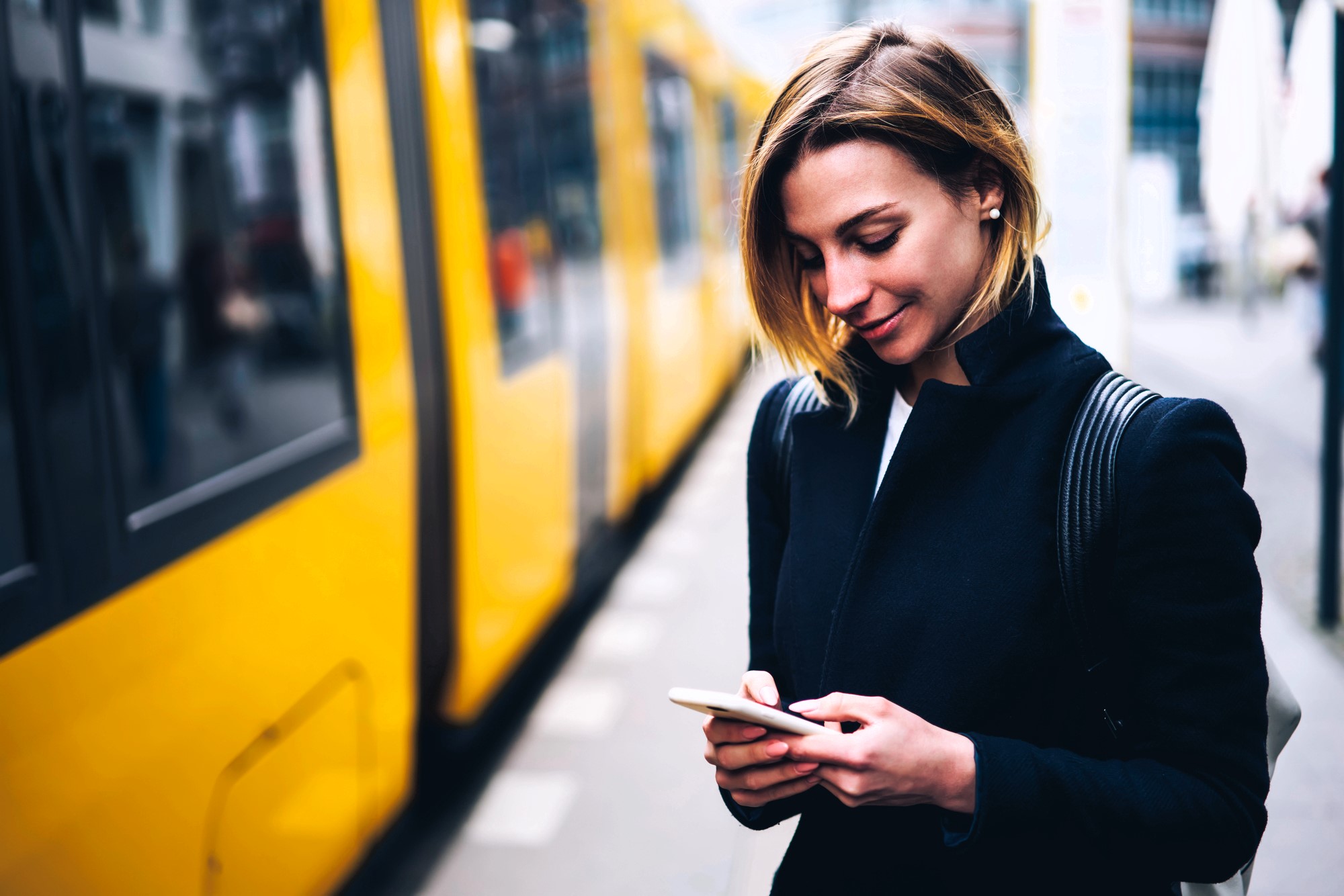 A woman with short hair is smiling while looking at her smartphone. She is standing next to a yellow train on a city street. She is wearing a black jacket and has a backpack. The background is slightly blurred.