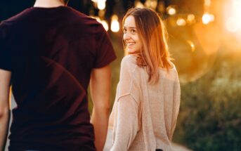 A woman smiles back at a man while walking together outdoors during sunset. The sun creates a warm glow around them. She is wearing a light sweater, and he is in a dark t-shirt. The scene is relaxed and cheerful.