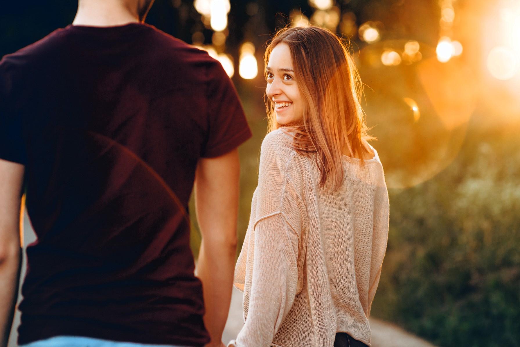 A woman smiles back at a man while walking together outdoors during sunset. The sun creates a warm glow around them. She is wearing a light sweater, and he is in a dark t-shirt. The scene is relaxed and cheerful.