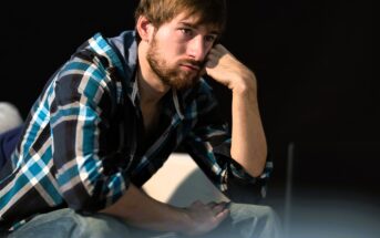 A man with a beard and brown hair, wearing a blue and black checkered shirt, sits with his head resting on his hand, gazing thoughtfully to the side. The background is dark, which contrasts with the light on his face.