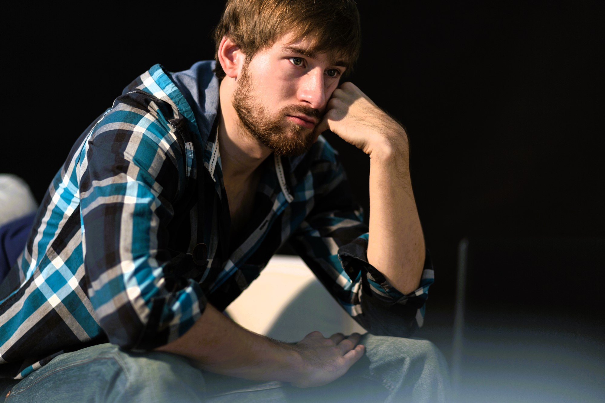 A man with a beard and brown hair, wearing a blue and black checkered shirt, sits with his head resting on his hand, gazing thoughtfully to the side. The background is dark, which contrasts with the light on his face.