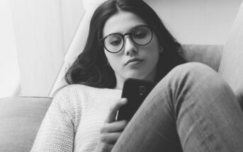 A woman with long hair and glasses is lying on a couch, looking intently at her smartphone. She is wearing a light sweater and jeans. The image is in black and white, creating a calm and focused atmosphere.