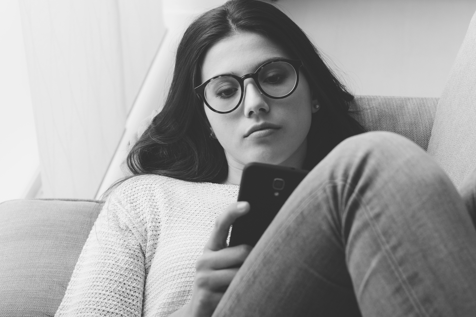 A woman with long hair and glasses is lying on a couch, looking intently at her smartphone. She is wearing a light sweater and jeans. The image is in black and white, creating a calm and focused atmosphere.