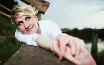 A person with short blonde hair and blue eyes, wearing a white shirt, lies on their stomach on a wooden surface outdoors. They gaze upward with a slight smile, with one hand reaching forward. The background includes blurred greenery and a fence.