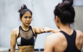 Woman with hair in a bun wearing a sports bra stands in front of a mirror, extending one arm forward. The background is blurred, focusing on her reflection and determined expression.