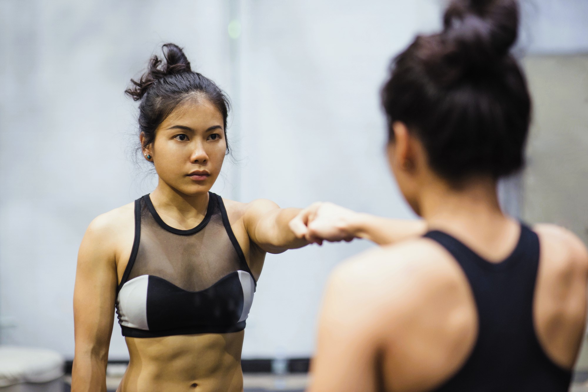 Woman with hair in a bun wearing a sports bra stands in front of a mirror, extending one arm forward. The background is blurred, focusing on her reflection and determined expression.