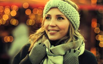 A woman smiles warmly while wearing a knitted headband, scarf, and gloves. The background features softly blurred, glowing lights, creating a festive and cozy atmosphere.