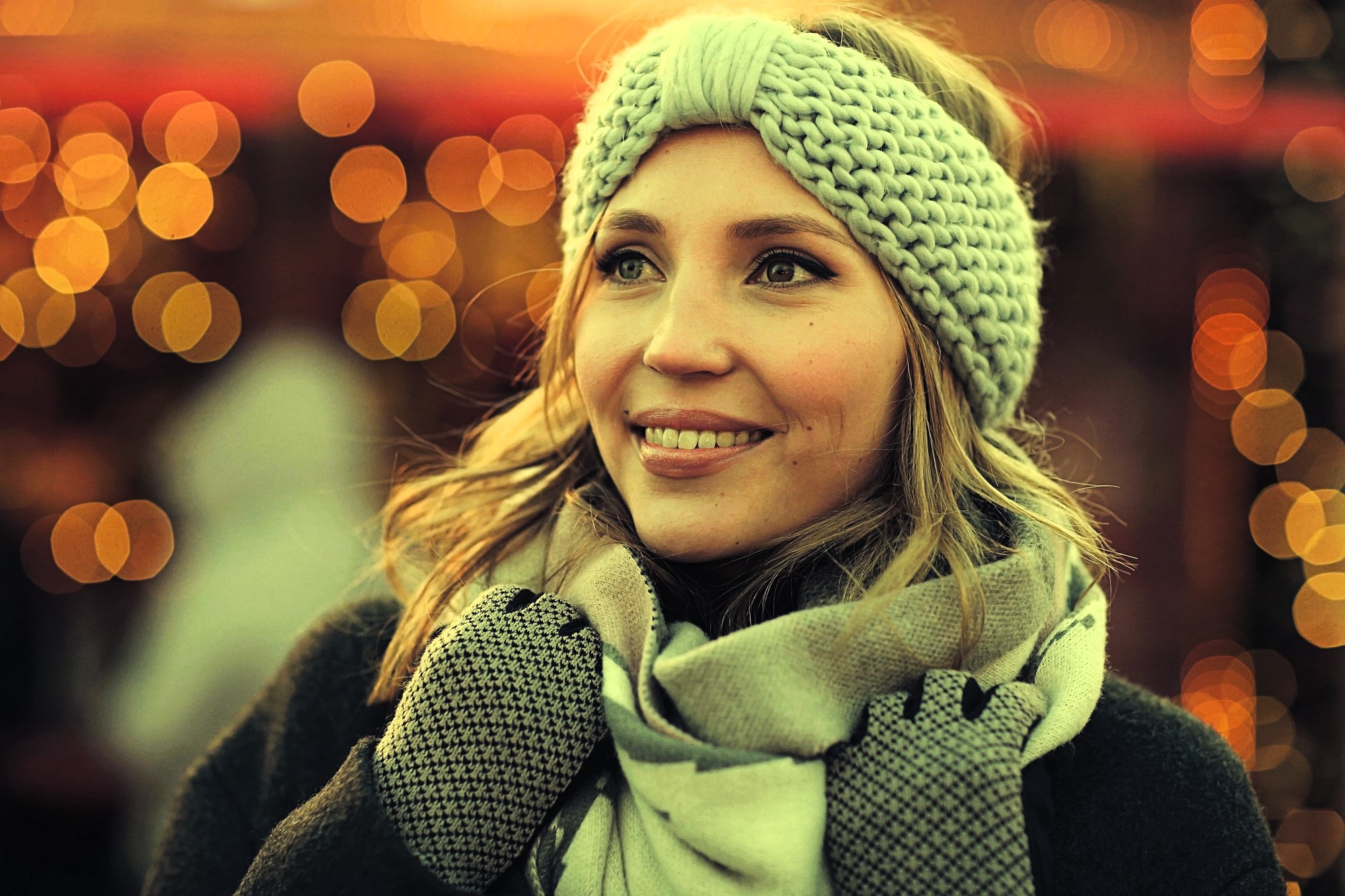 A woman smiles warmly while wearing a knitted headband, scarf, and gloves. The background features softly blurred, glowing lights, creating a festive and cozy atmosphere.