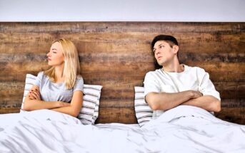 A man and a woman sit in bed with crossed arms, looking away from each other, appearing upset or in disagreement. They are sitting against a wooden headboard, wearing casual clothes. The bed is covered with a white blanket and striped pillows.