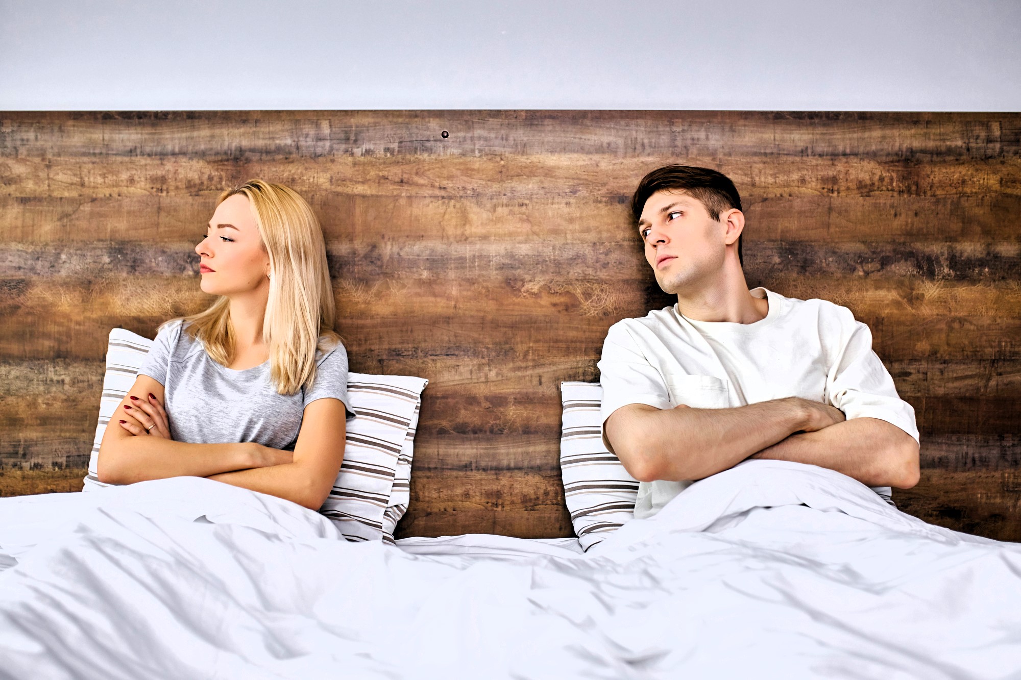 A man and a woman sit in bed with crossed arms, looking away from each other, appearing upset or in disagreement. They are sitting against a wooden headboard, wearing casual clothes. The bed is covered with a white blanket and striped pillows.