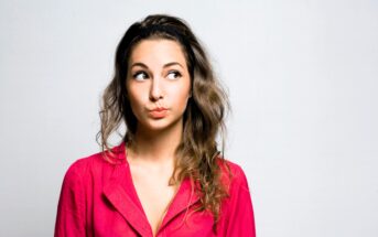 A woman with long brown hair, wearing a bright pink blouse, looks up with a thoughtful expression. She stands against a plain gray background.