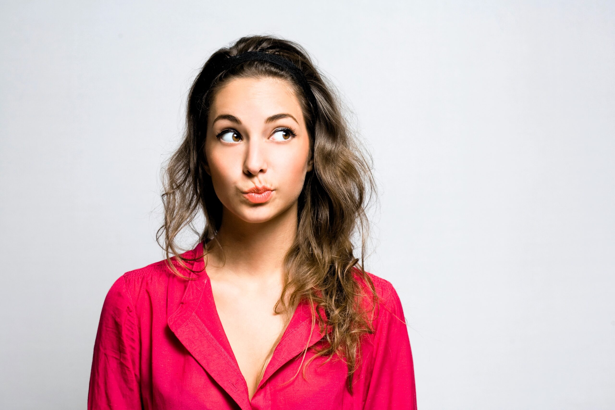 A woman with long brown hair, wearing a bright pink blouse, looks up with a thoughtful expression. She stands against a plain gray background.