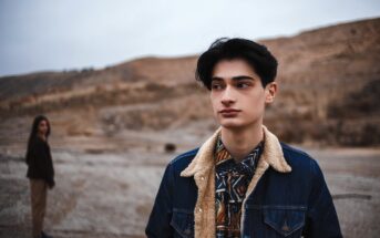 A young person in a denim jacket with a shearling collar stands in focus, looking to the side, in a barren, hilly landscape. In the background, another figure in casual attire stands out of focus. The sky appears overcast.