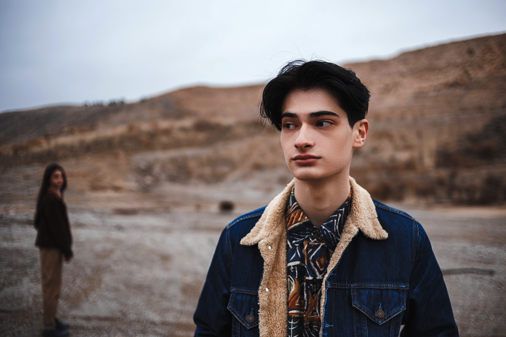 A young person in a denim jacket with a shearling collar stands in focus, looking to the side, in a barren, hilly landscape. In the background, another figure in casual attire stands out of focus. The sky appears overcast.