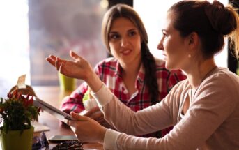 Two women sit at a table in a cafe, engaged in a conversation. One holds a tablet, gesturing with her hand, while the other listens attentively. A slice of cake and a small plant are on the table. The background is softly blurred.