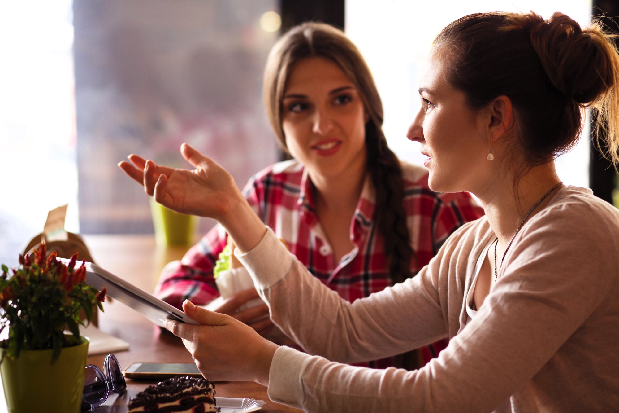 Two women sit at a table in a cafe, engaged in a conversation. One holds a tablet, gesturing with her hand, while the other listens attentively. A slice of cake and a small plant are on the table. The background is softly blurred.