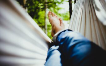 Close-up of a person's feet as they relax in a white hammock. They are wearing blue jeans, and the surrounding area is filled with lush green foliage, suggesting a serene outdoor setting. The image conveys a sense of calm and leisure.