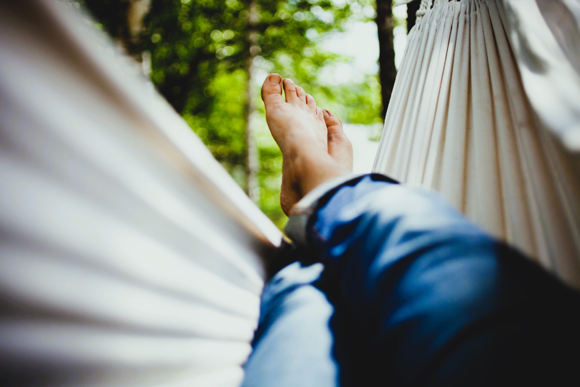 Close-up of a person's feet as they relax in a white hammock. They are wearing blue jeans, and the surrounding area is filled with lush green foliage, suggesting a serene outdoor setting. The image conveys a sense of calm and leisure.