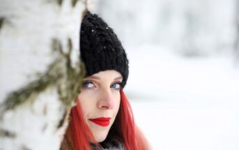 A woman with bright red hair and red lipstick is wearing a black knit hat and gray scarf, partially obscured behind a snow-covered tree. The background is a snowy, blurred winter landscape.