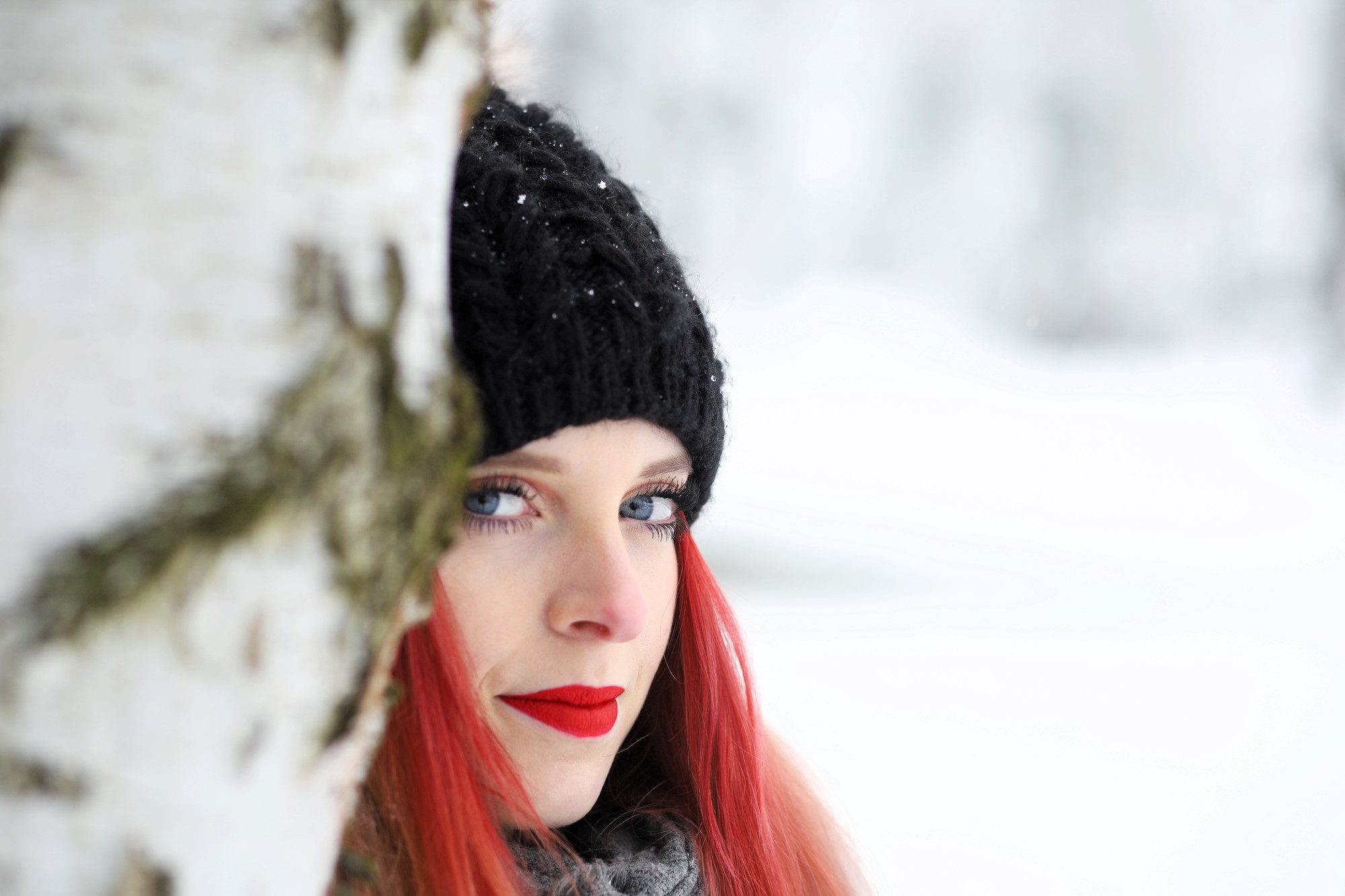 A woman with bright red hair and red lipstick is wearing a black knit hat and gray scarf, partially obscured behind a snow-covered tree. The background is a snowy, blurred winter landscape.