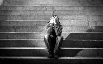 A person sits on stone steps with their head in their hands, appearing distressed. The image is in black and white, emphasizing the texture of the steps and the individual's clothing. The setting seems isolated and somber.