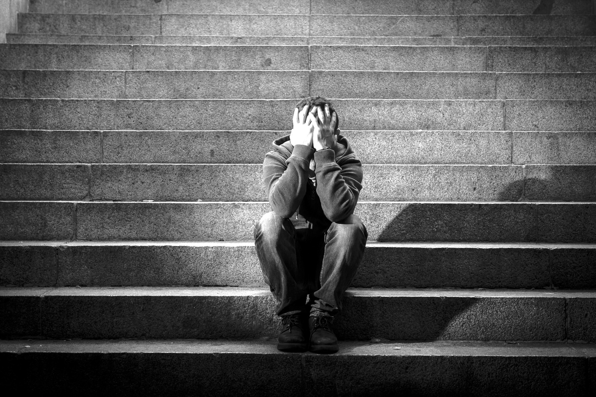 A person sits on stone steps with their head in their hands, appearing distressed. The image is in black and white, emphasizing the texture of the steps and the individual's clothing. The setting seems isolated and somber.