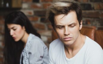 A man with a white t-shirt and styled hair looks down pensively while sitting on a brown chair. A woman with long dark hair, wearing a striped shirt, sits blurred in the background against a brick wall.