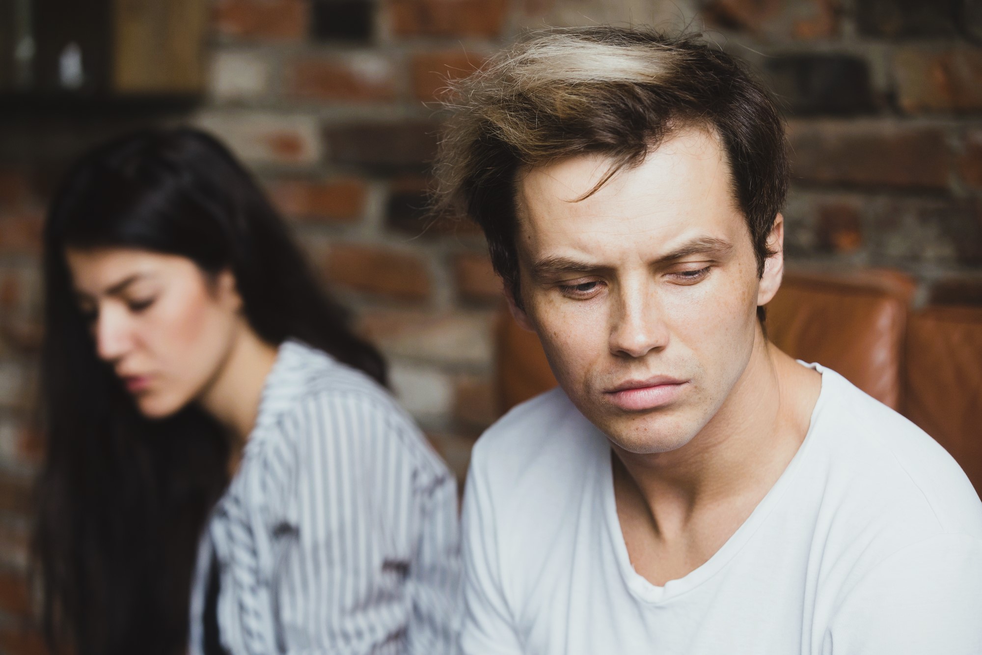 A man with a white t-shirt and styled hair looks down pensively while sitting on a brown chair. A woman with long dark hair, wearing a striped shirt, sits blurred in the background against a brick wall.