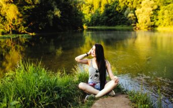 A woman with long dark hair sits cross-legged on a dirt path by a calm lake, surrounded by lush green grass and trees. She is wearing a gray tank top and shorts, with sunglasses resting on her chest, enjoying the serene nature setting.