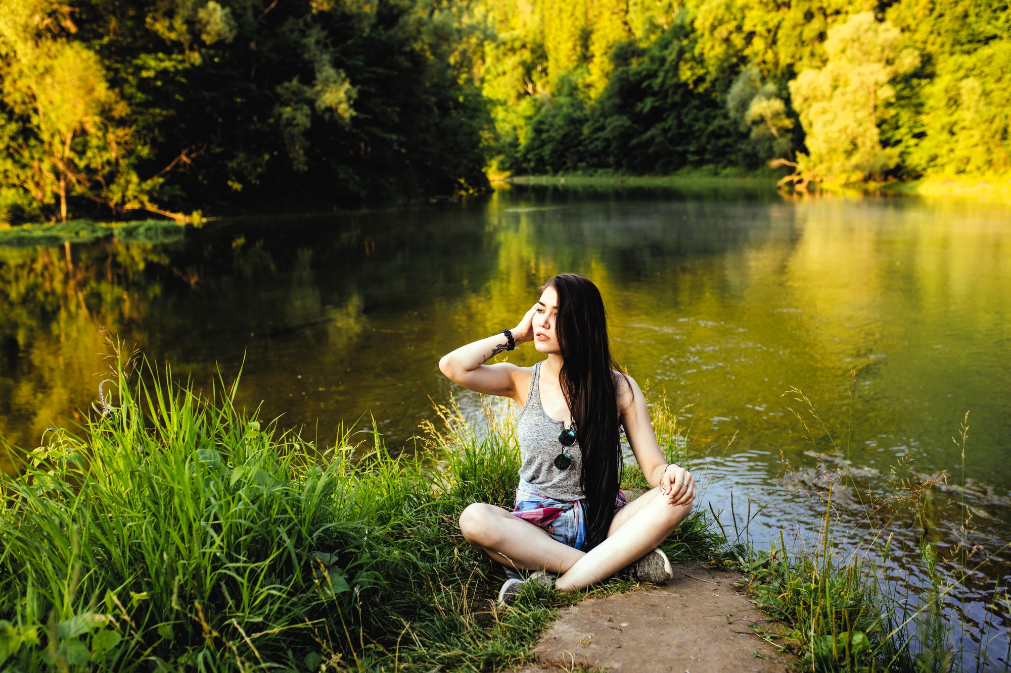 A woman with long dark hair sits cross-legged on a dirt path by a calm lake, surrounded by lush green grass and trees. She is wearing a gray tank top and shorts, with sunglasses resting on her chest, enjoying the serene nature setting.