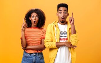 A surprised-looking man and woman stand against a bright orange background. The woman has curly hair and wears a striped orange top and jeans, while the man has short hair and wears a yellow jacket over a white shirt. Both have their index fingers raised.