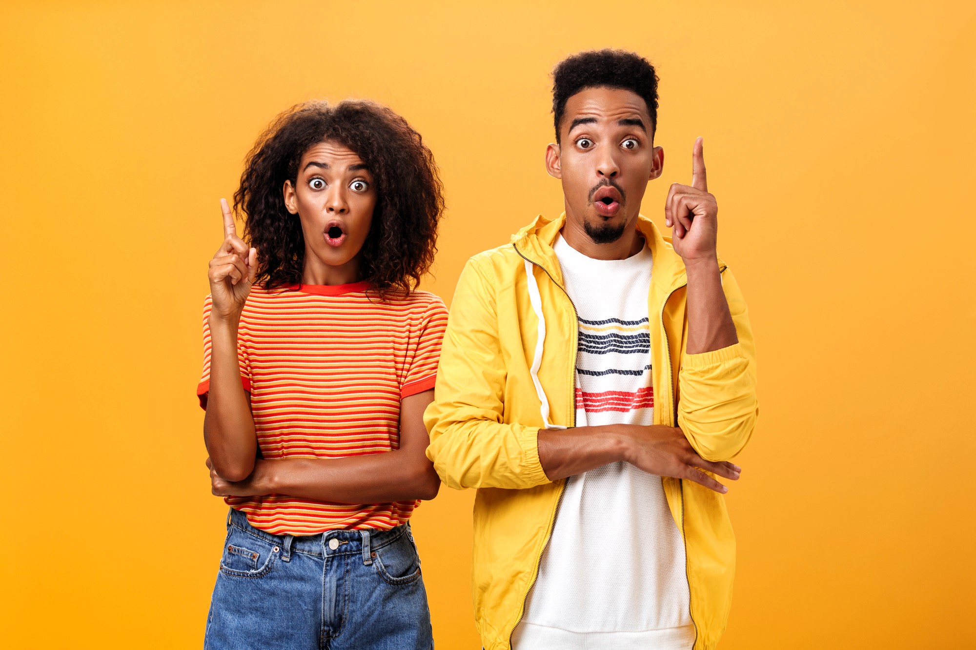 A surprised-looking man and woman stand against a bright orange background. The woman has curly hair and wears a striped orange top and jeans, while the man has short hair and wears a yellow jacket over a white shirt. Both have their index fingers raised.