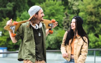 A man and woman outdoors, leaning against a railing. The man, wearing a cap and headphones, holds a skateboard on his shoulders. The woman, wearing a cap and denim jacket, smiles at him. Greenery is visible in the background.