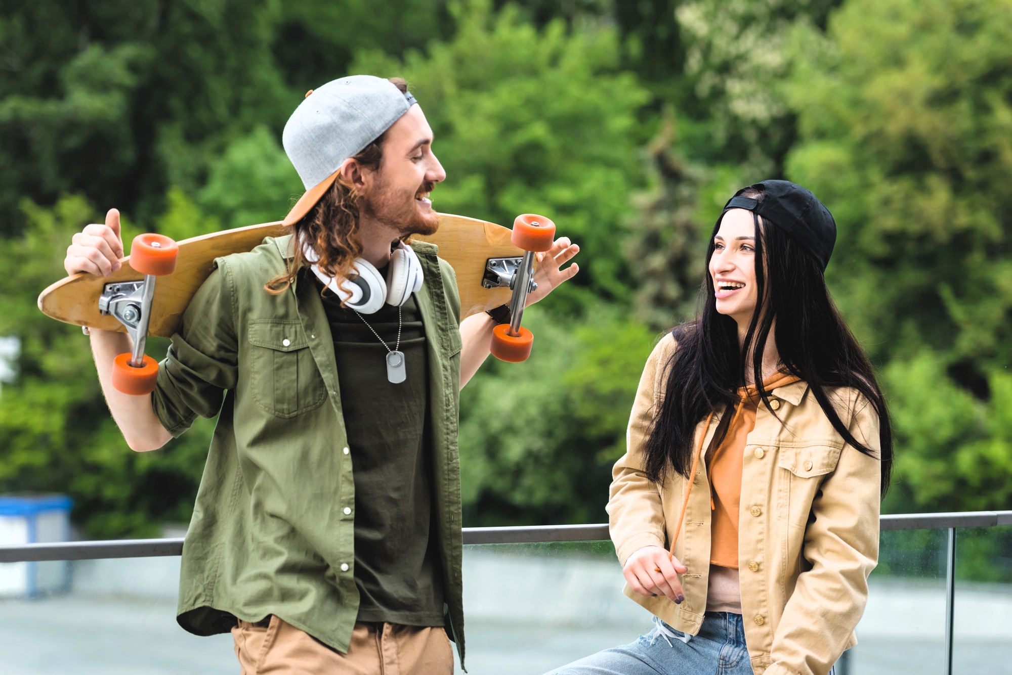 A man and woman outdoors, leaning against a railing. The man, wearing a cap and headphones, holds a skateboard on his shoulders. The woman, wearing a cap and denim jacket, smiles at him. Greenery is visible in the background.