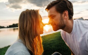 A couple sits close on a grassy area by a lake at sunset, looking into each other's eyes. The sun sets behind them, creating a warm glow in the sky. Both are smiling softly, enjoying the serene moment.