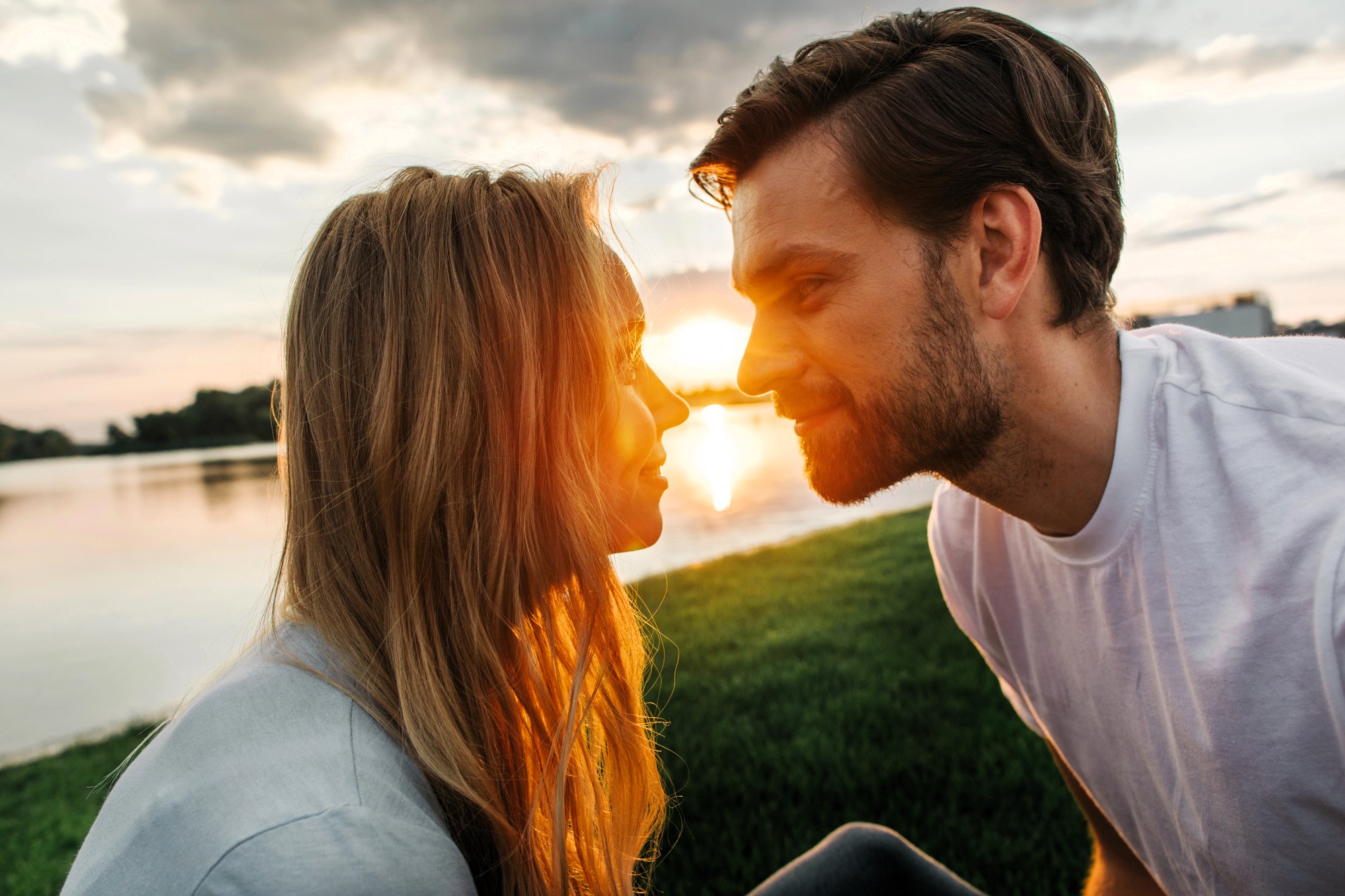 A couple sits close on a grassy area by a lake at sunset, looking into each other's eyes. The sun sets behind them, creating a warm glow in the sky. Both are smiling softly, enjoying the serene moment.