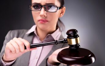 A woman in a gray suit and glasses holds a wooden gavel, poised to strike. She has a serious expression and stands against a plain dark background, suggesting a legal or judicial context.