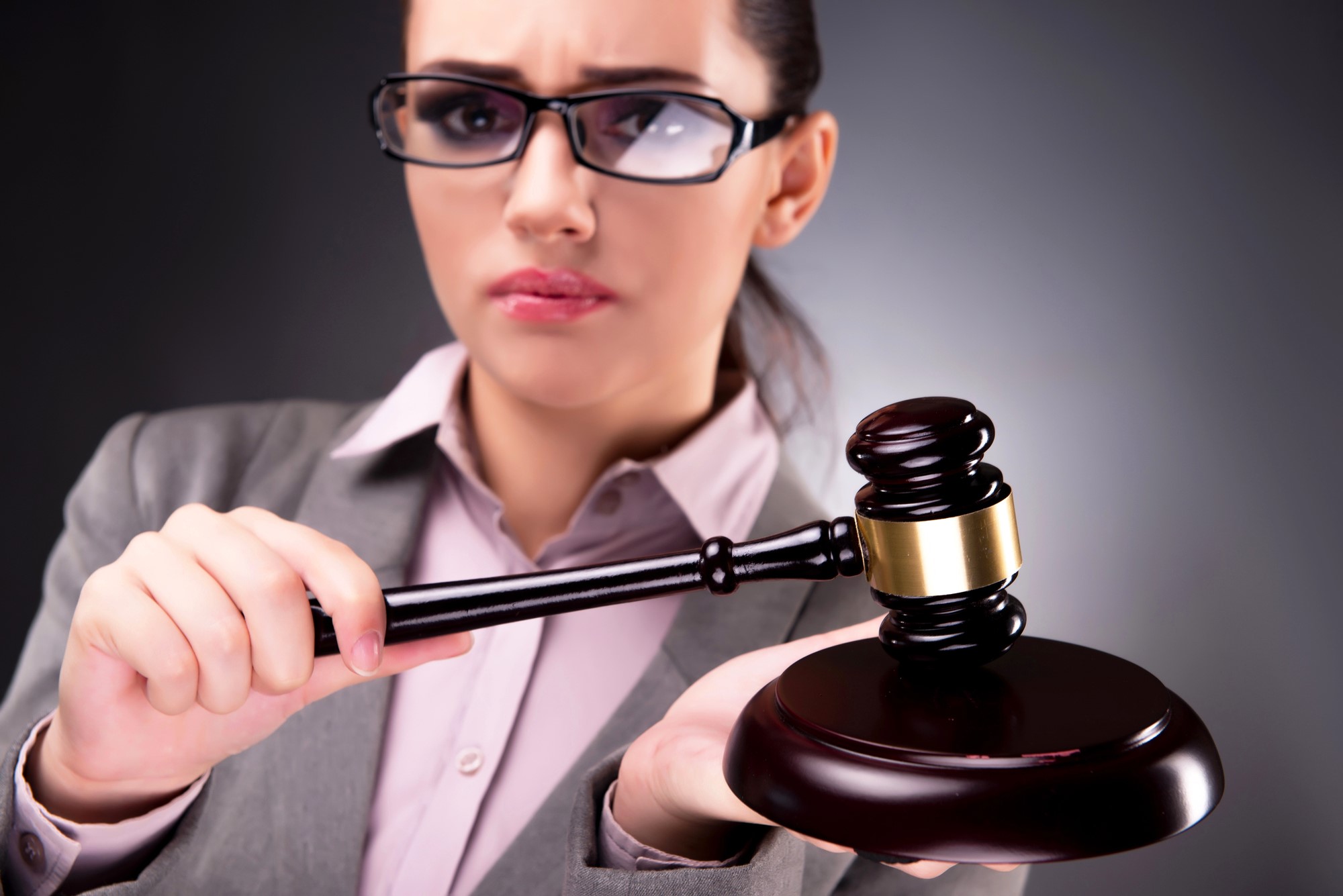 A woman in a gray suit and glasses holds a wooden gavel, poised to strike. She has a serious expression and stands against a plain dark background, suggesting a legal or judicial context.