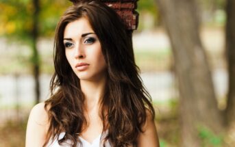 Woman with long brown hair and a contemplative expression standing outdoors, leaning against a brick pillar. She is wearing a white top, and the background is blurred with trees and greenery.